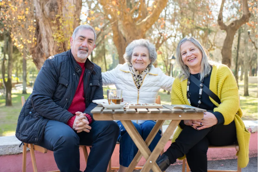 Three older friends smiling together at a table in a park